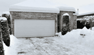 Snow-covered brick house with a garage and trimmed bushes in winter.